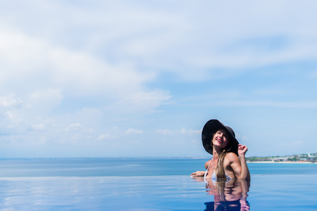 Young woman in white bikini and grey hat enjoying a sun in the infinity pool. Vacations and summer conceptの写真素材