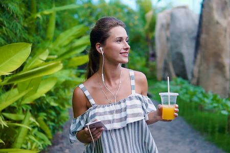 technology and healthy lifestyle concept - woman in earphones with takeaway smoothie drink or shake or juice in plastic cup and smartphone listening to music at summer parkの写真素材