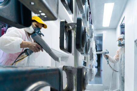 Powder coating of metal parts. Man and woman in protective suit sprays white powder paint from gun on metal productsの写真素材