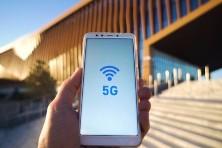 man hand holding a smartphone and 5g signal symbol on screen with broadcast antenna icon on city...