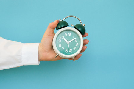 Hand holding a classic alarm clock on a blue backgroundの写真素材