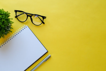 Flat lay with a blank spiral note, glasses, pen and home plant on yellow background