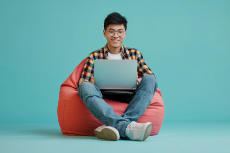 Full body young happy young man of Asian ethnicity sit in bag chair use work on laptop isolated on plain pastel background. AI generativeの素材