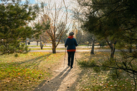 A full-length portrait of a Caucasian woman wearing a jacket and hat with Nordic walking poles walks through a pine forest in a city park, back viewの写真素材