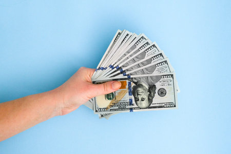 A woman's hand holds a few hundred-dollar bill on a blue background