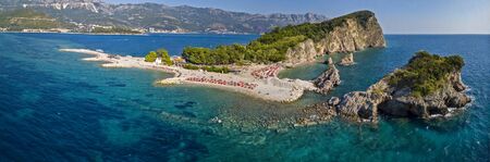 Aerial view of Sveti Nikola, Budva island, Montenegro. Hawaii beach, umbrellas and bathers and crystal clear waters. Jagged coasts with sheer cliffs overlooking the transparent sea. Wild nature and Mediterranean maquisの写真素材