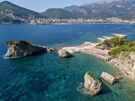 Aerial view of Sveti Nikola, Budva island, Montenegro. Hawaii beach, umbrellas and bathers and crystal clear waters. Jagged coasts with sheer cliffs overlooking the transparent sea. Wild nature and Mediterranean maquisの写真素材