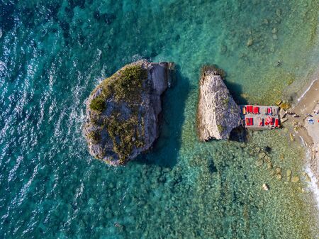 Aerial view of Sveti Nikola, Budva island, Montenegro. Hawaii beach, umbrellas and bathers and crystal clear waters. Jagged coasts with sheer cliffs overlooking the transparent sea. Wild nature and Mediterranean maquisの写真素材