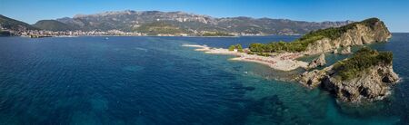 Aerial view of Sveti Nikola, Budva island, Montenegro. Hawaii beach, umbrellas and bathers and crystal clear waters. Jagged coasts with sheer cliffs overlooking the transparent sea. Wild nature and Mediterranean maquisの写真素材