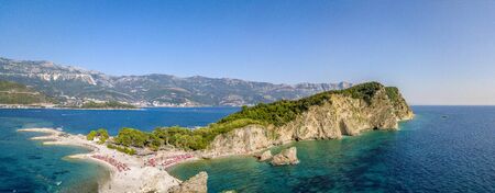 Aerial view of Sveti Nikola, Budva island, Montenegro. Hawaii beach, umbrellas and bathers and crystal clear waters. Jagged coasts with sheer cliffs overlooking the transparent sea. Wild nature and Mediterranean maquisの写真素材
