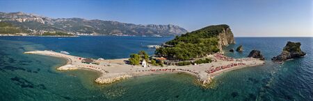 Aerial view of Sveti Nikola, Budva island, Montenegro. Hawaii beach, umbrellas and bathers and crystal clear waters. Jagged coasts with sheer cliffs overlooking the transparent sea. Wild nature and Mediterranean maquisの写真素材