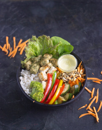 Pesto chicken barley salad bowl with broccoli, bell pepper, zucchini, and fresh lettuce served in a black bowl with dressing on the side.の写真素材