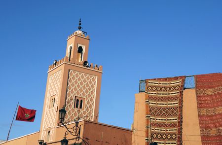 Moroccan Carpets for sale at a shop and minaret of a mosque with Moroccan national flag in Marrakesh
