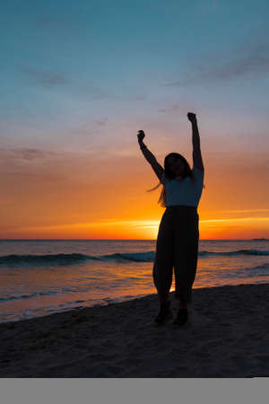 a silhouette of a young girl with her arms up in backlight at sunset on the beachの写真素材
