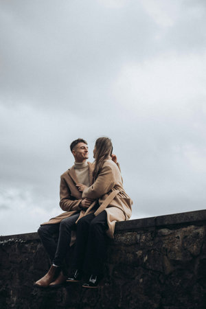 stylish man and woman in beige coats sitting on wall and looking away