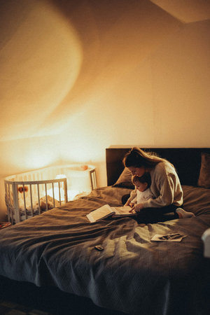 Young woman reading book while sitting on bed in bedroom at home.