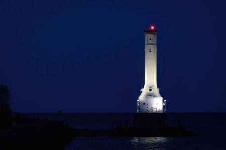 A striking night scene featuring a tall white lighthouse illuminated against the deep blue sky, standing along the shoreline with its red beacon glowing.の写真素材