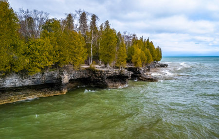 View from the water showing the rugged limestone cliffs and rocky shoreline of Cave Point County Park in Wisconsin with waves and forest backdrop.の写真素材