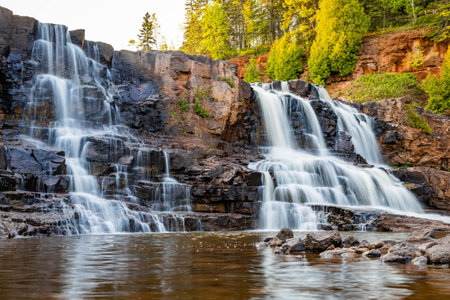 A scenic view of Middle Falls at Gooseberry Falls State Park during a golden sunset, showcasing the beauty of the cascading waterの写真素材