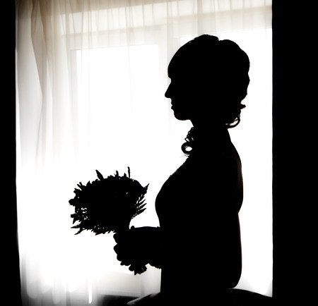 Black silhouette of beautiful bride with posy against white windowの写真素材