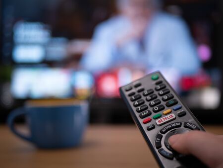 UK, Jan 2020: close up of TV remote with television behind showing Netflix menu documentary