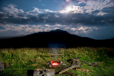 Campfire on green lawn near mountain at night with dark sky and moon lightの写真素材