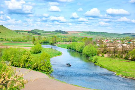 Rural view to river banks with farm, field, green trees and blue cloudy sky