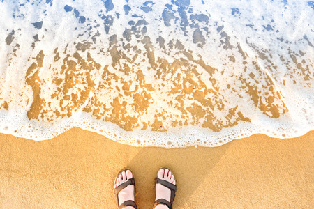 Woman's feet in sandals on yellow beach sand with blue sea wave and white foamの写真素材