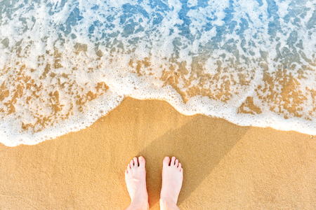 Woman's feet on yellow beach sand with sea wave and white foamの写真素材