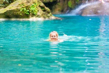 Red haired young woman swims in emerald blue water of tropical lake. Erawan National park, Kanchanaburi, Thailandの写真素材