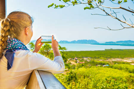 Young woman with mobile phone taking image of Thailand mountain island sea landscapeの写真素材