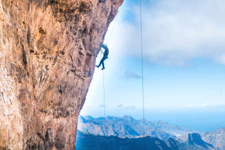 Rock climber climbing up overhanging cliff with Canary Island mountain range and sea on backgroundの写真素材