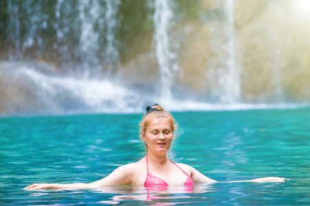 Red haired young woman in pink bikini swimsuit relaxes in emerald tropical lake with waterfall. Erawan National park, Kanchanaburi, Thailandの写真素材
