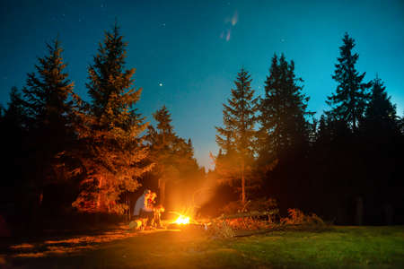 Bonfire in night forest and people near fire under night dark sky with stars