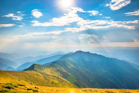 Mountains landscape with peaks, sun, clouds and grass fieldの写真素材