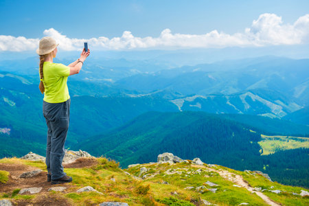 Young woman standing on mountain cliff and taking travel selfie on smartphoneの写真素材