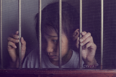 sad asian child standing behind the wire screen window in split tone.  Unhappy kid alone at home. Upset.の写真素材