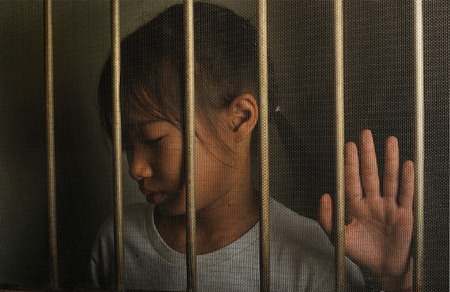 sad asian child standing behind the wire screen window.  Unhappy kid  alone at home. Upset in the dark room.の写真素材