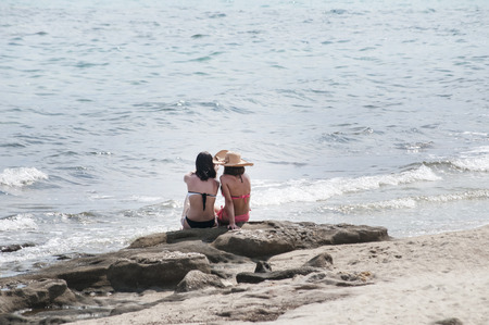 Two girls sitting back on sea beach rockの写真素材