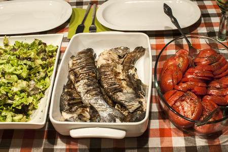 Home table stacked with white porcelain dishes with grilled trout, lettuce and sliced tomatoesの写真素材