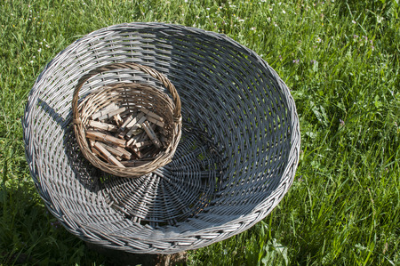 Big old weathered empty wicker laundry basket and small basket full of wooden clothes pegs on wooden log on green grass country garden meadowの写真素材