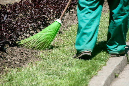 Maintenance worker in park garden cleans the flower beds with plastic garden broom closeup