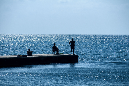 Stone quay in sea bay of calm blue sea waters and three human silhouettes in clear sunny summer dayの写真素材