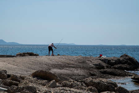 Landscape with man fishing with fishing rods on sea rock in sunny summer dayの写真素材