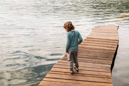 A boy stands on a wooden pier and looks into the distance. Little boy on wooden boardwalk leading...