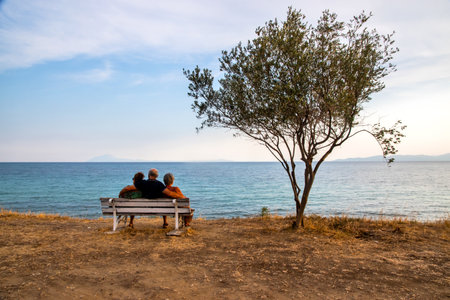 Idyllic landscape with people sitting on bench near tree in the background of a sea bay at sunsetの写真素材