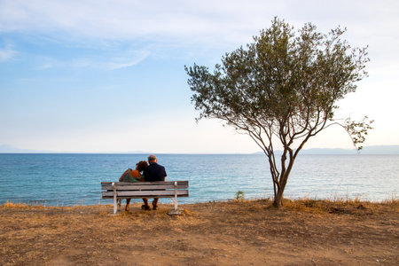 Idyllic landscape with people sitting on bench near tree in the background of a sea bay at sunsetの写真素材