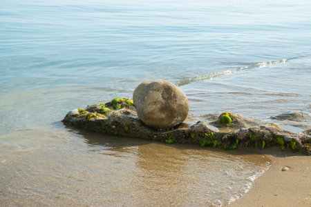 A smooth round stone resting on an algae-covered sea rock by a calm, sandy shoreの写真素材