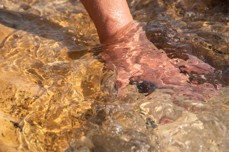 Close-up of a woman's hand touching a warm, smooth beach stone washed by clear, flowing seawater in...