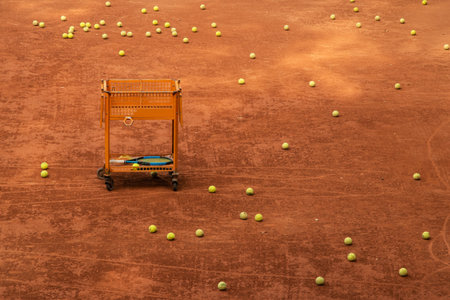 Coaching cart filled with tennis balls and rackets placed on clay court, emphasizing structured learning and sports preparationの写真素材